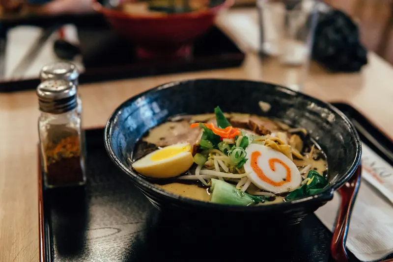 Reference photograph of ramen with egg, sliced pork, greens and a spiral fish cake in a black bowl